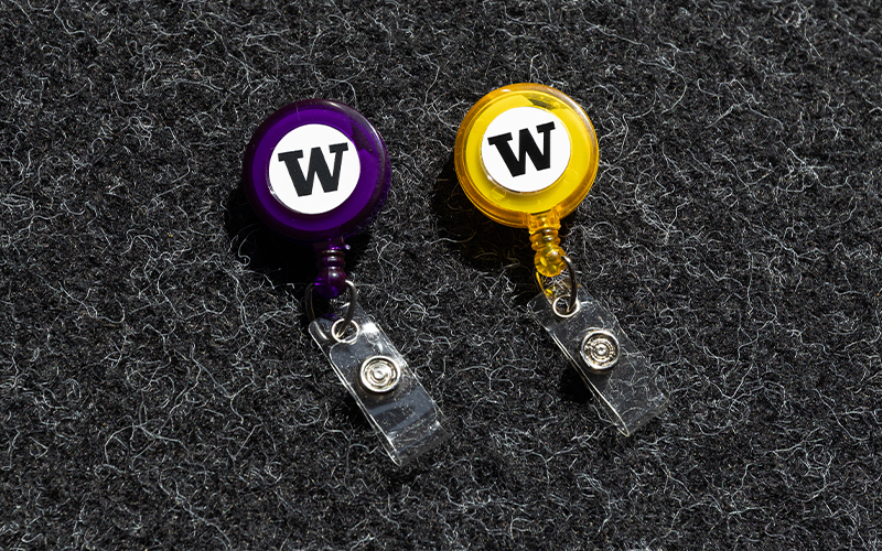 Purple and yellow badge reels with the UW logo, each with a plastic clasp and rolled up stretchy string to extend or retract the badge.