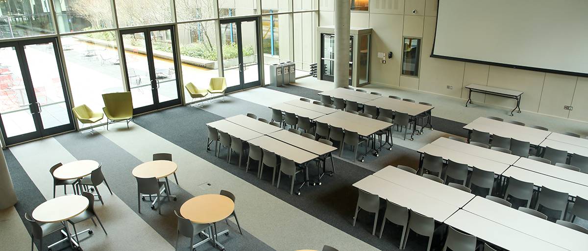 A mixed use meeting space with several large tables and seating for eighty in front of a large projector screen.