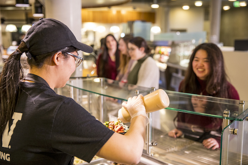 A UW Dining employee pours sauce onto a bowl while a student watches smiling.